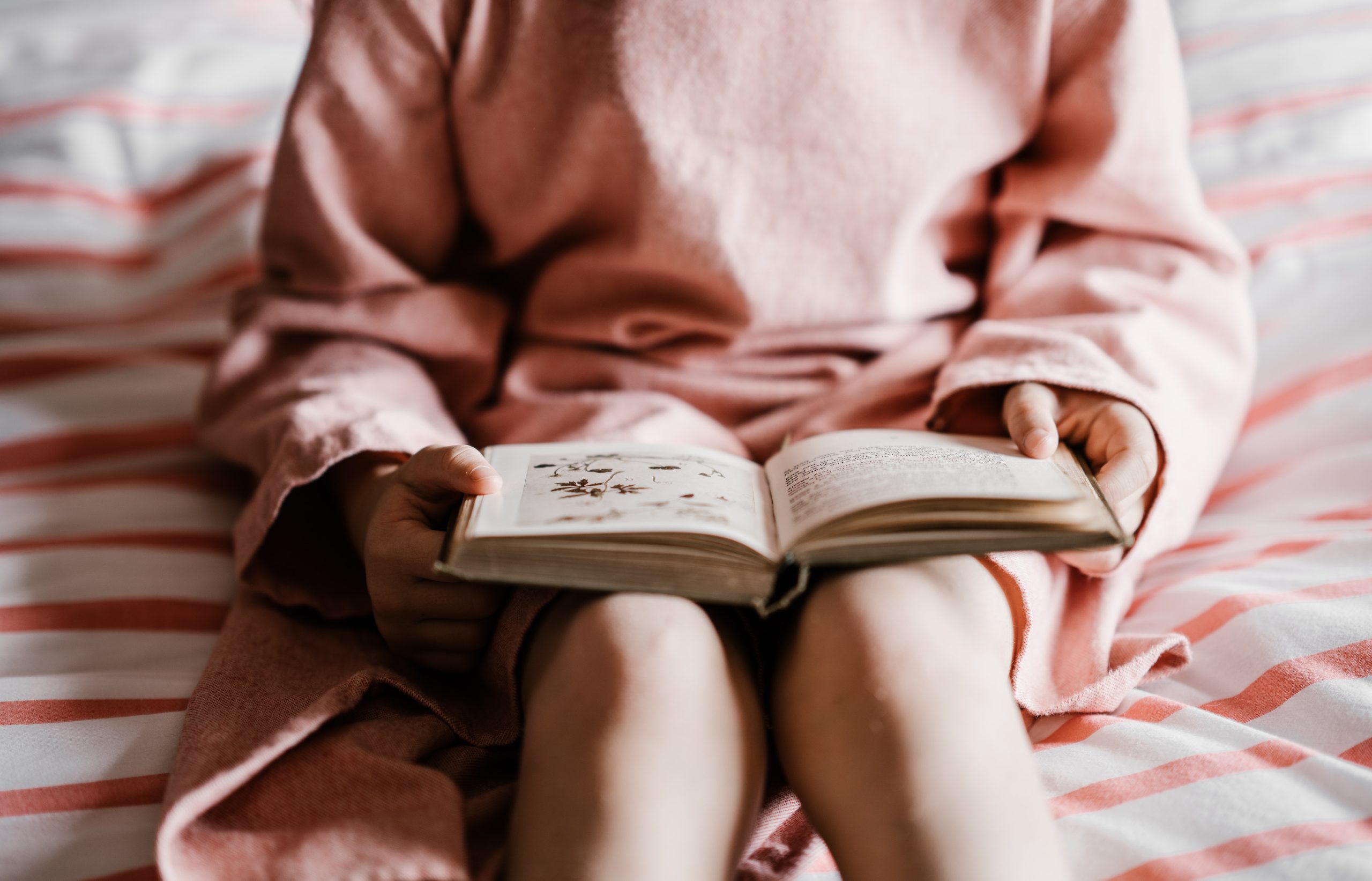 Girl in pink dress reading a book on her bed