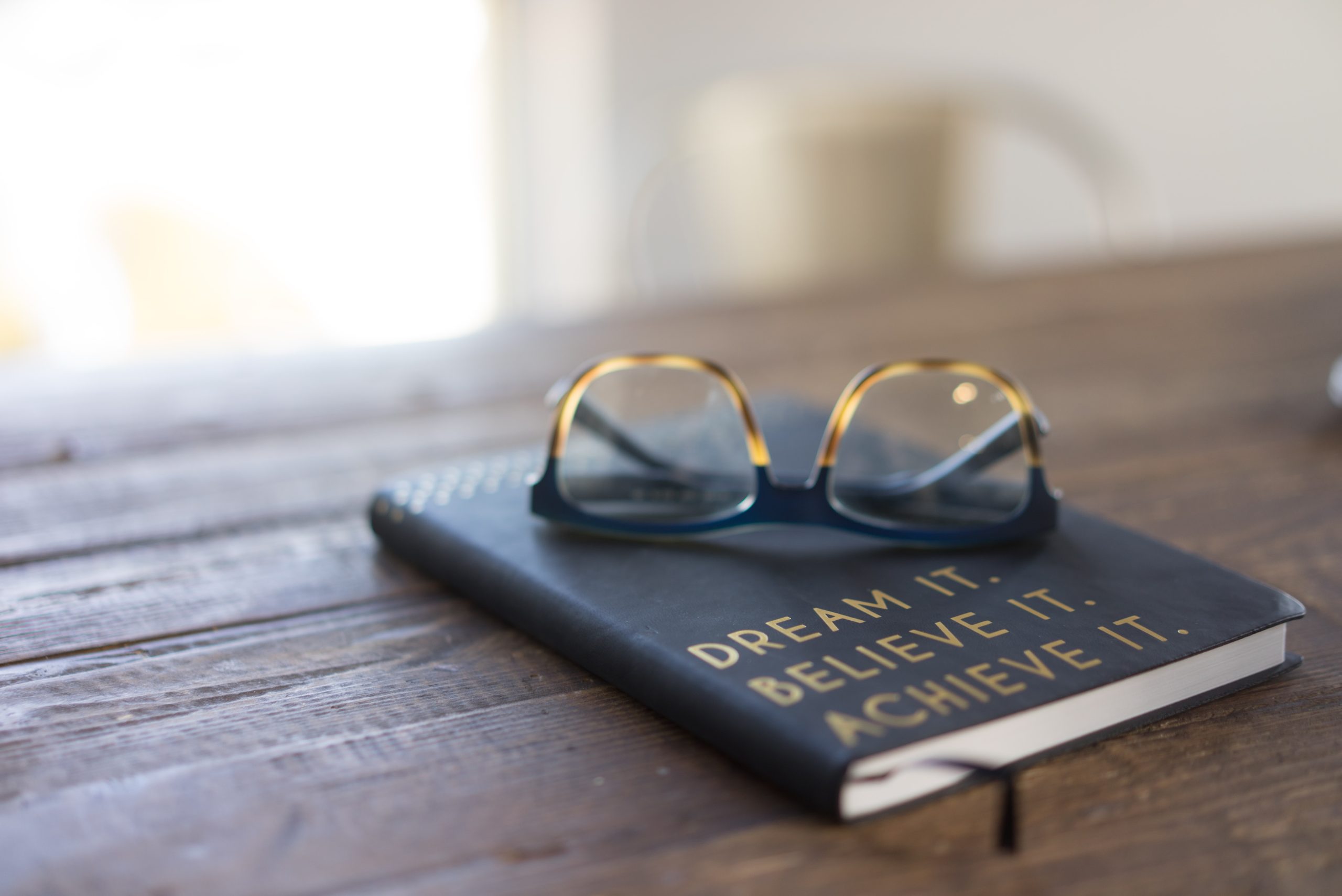 Book with text that reads ‘dream it, believe it, achieve it’ and glasses on top of the book 