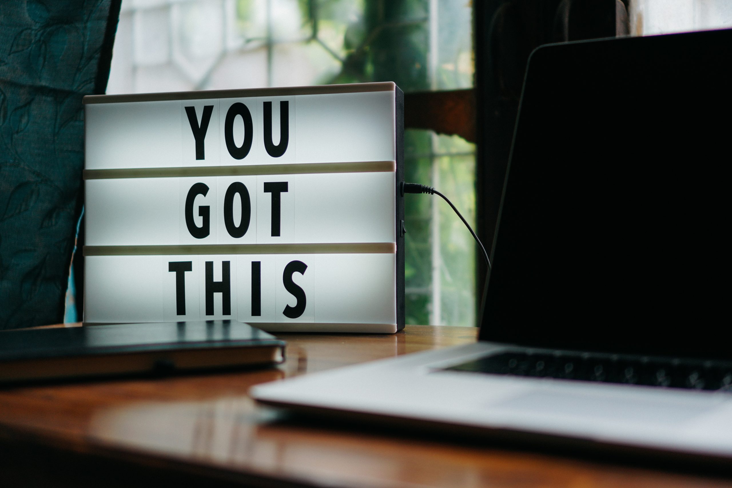 A desktop beside a window with a laptop, a book and a lit alphabetical display that reads 'You got this'.