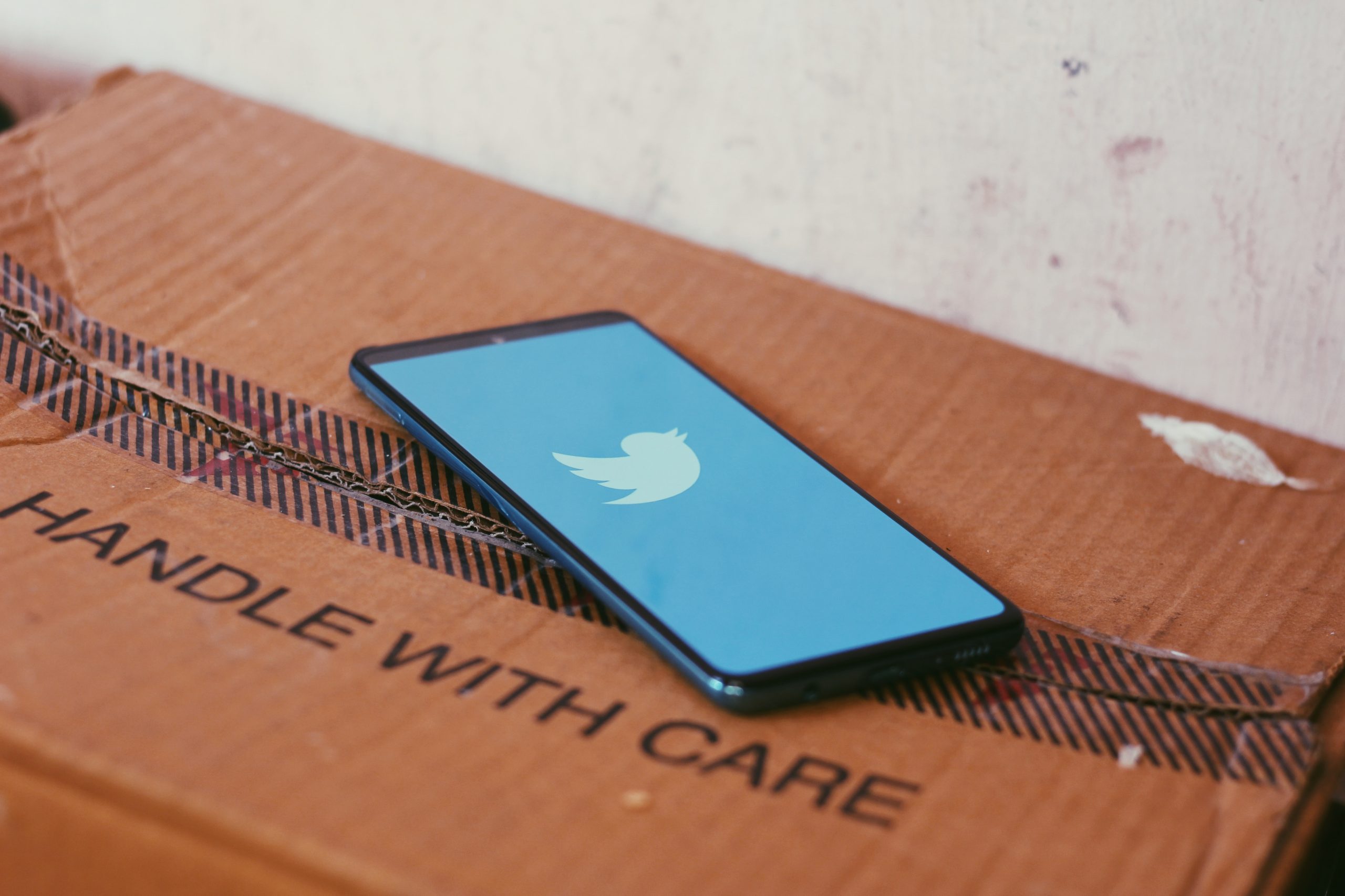 A smartphone lying on a carton that reads ‘Handle with care’ while the phone’s screen has twitter’s symbol.